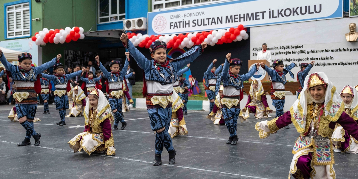 İzmir Bornova Fatih Sultan Mehmet İlkokulu'nun bahçesinde, geleneksel halk oyunları kıyafetleri giymiş bir grup öğrenci gösteri yapıyor. Mavi işlemeli yelek ve şalvar giyen erkek öğrenciler ile altın sarısı detaylı, işlemeli kıyafetler ve başlıklar takan kız öğrenciler, okul binasının önündeki tören alanında dans ediyor. Arka planda kırmızı ve beyaz balonlarla süslenmiş okul binası ve duvarda Atatürk büstü ile okulun ismi yer alıyor.