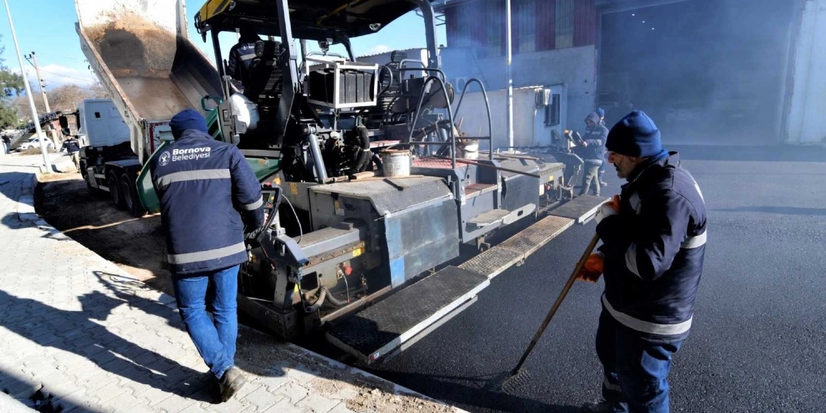 Bornova Belediyesi ekipleri, bir asfalt serim makinesi ve kamyon kullanarak yol yenileme çalışması yapıyor. İşçilerden biri makineyi kontrol ederken, diğeri elindeki tırmıkla yeni dökülen asfaltı düzeltiyor.