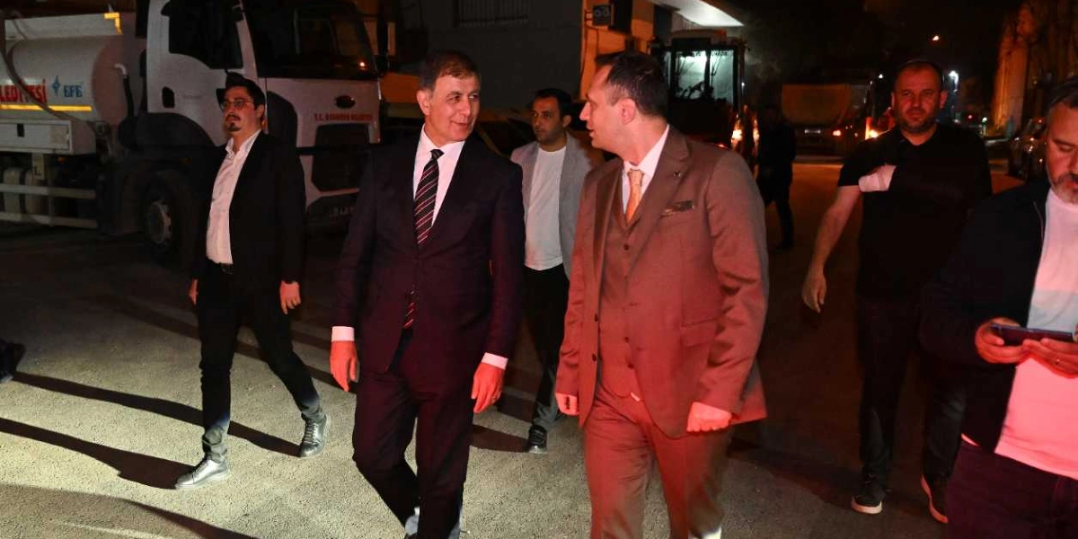 Gece vakti dış mekanda, takım elbiseli iki erkek yan yana yürürken birbirleriyle sohbet ediyor. Sol tarafta koyu renk takım elbiseli ve çizgili kravatlı bir kişi, sağ tarafta ise kahverengi yelekli takım elbiseli başka bir kişi yer alıyor. Arka planda belediyeye ait bir kamyon ve çevrelerinde yürüyen diğer kişiler bulunuyor.
