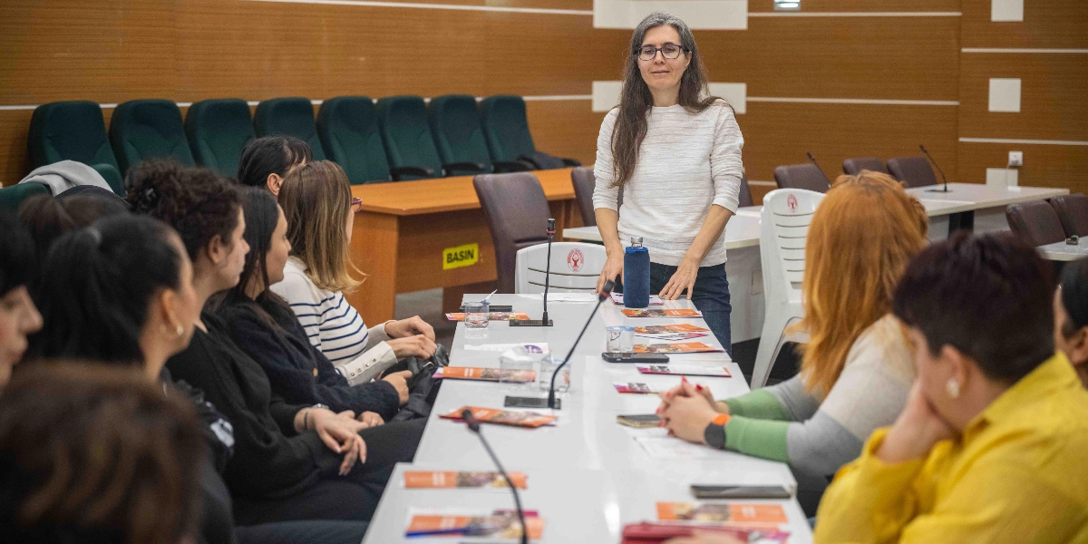 Ahşap panelli bir toplantı salonunda, uzun bir masanın başında ayakta duran gözlüklü ve uzun saçlı bir kadın, masanın etrafında oturan bir grup kadına hitap ediyor. Masanın üzerinde mikrofonlar, su şişesi ve üzerinde görseller bulunan broşürler yer alıyor. Katılımcılar dikkatle konuşmacıyı dinliyor.