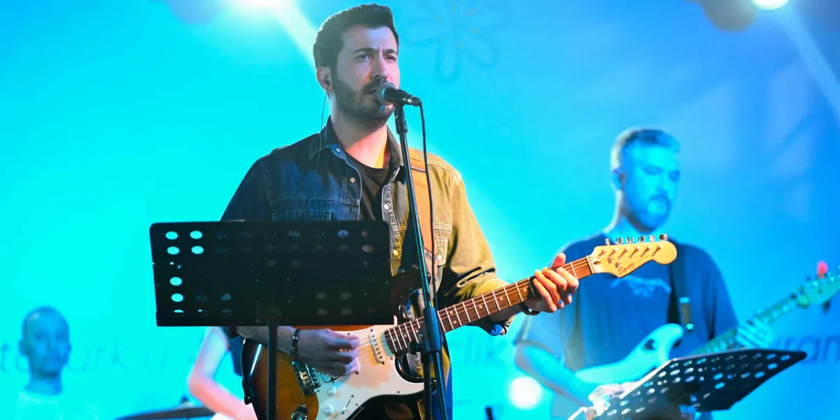 Mavi ışıklarla aydınlatılmış bir sahnede, elinde elektro gitarıyla şarkı söyleyen sakallı bir erkek solist ve arkasında gitar çalan grup arkadaşları yer alıyor.