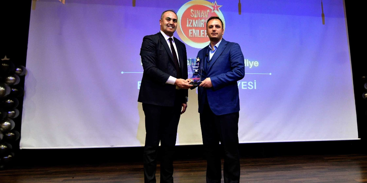Koyu renk takım elbiseli iki erkek, sahne üzerinde birbirlerine bir ödül plaketi takdim ederken gülümseyerek poz veriyor. Arka planda üzerinde 