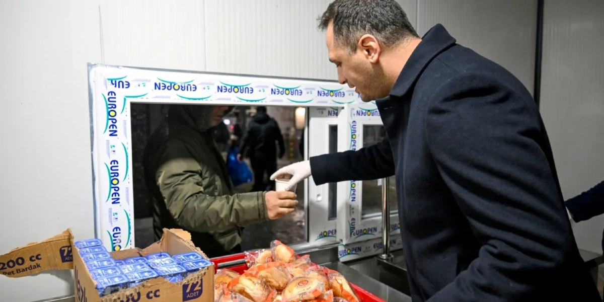 Koyu renkli palto giymiş bir erkek, beyaz eldivenli eliyle bir pencereden dışarıdaki kişiye içinde sıcak içecek bulunan bir karton bardak uzatıyor. Pencerenin önündeki tezgahta, içinde paketli sandviçlerin bulunduğu kırmızı bir kasa ve yanında 200 cc'lik bardakların olduğu bir karton kutu yer alıyor.