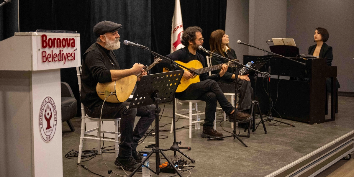 Bornova Belediyesi kürsüsünün yanında, sahnede canlı müzik icra eden dört kişilik bir grup yer alıyor. En solda kasketli, beyaz sakallı bir müzisyen bağlama çalıyor; yanında gitar çalan bir erkek, ayakta şarkı söyleyen bir kadın ve en sağda piyano başında oturan bir kadın müzisyen bulunuyor. Müzisyenlerin önünde mikrofonlar ve nota sehpaları yer alıyor, arka planda ise Bornova Belediyesi bayrağı asılı duruyor.