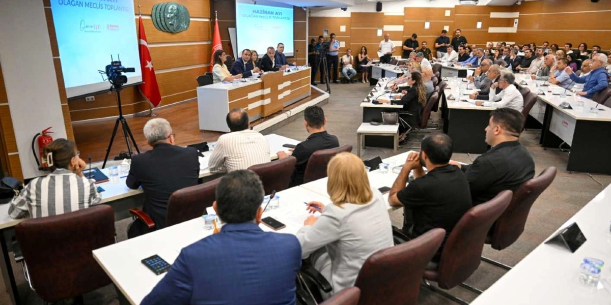 Bornova Belediyesi meclis salonunda düzenlenen Haziran ayı olağan meclis toplantısı. Salonun ön kısmındaki kürsüde yetkililer otururken, izleyiciler ve meclis üyeleri sıralar halinde oturarak toplantıyı takip ediyor. Salonun duvarında Türk bayrağı ve Atatürk rölyefi bulunurken, ön taraftaki projeksiyon ekranlarında toplantı başlığı yer alıyor.