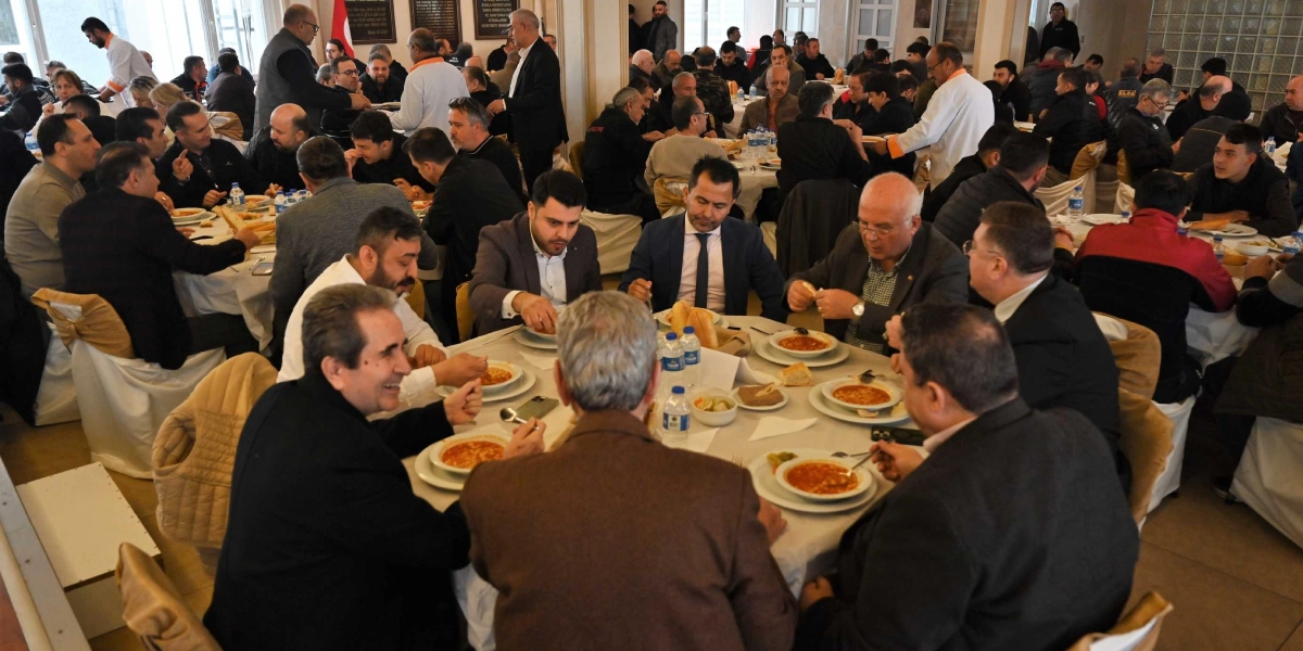 Geniş bir salonda, beyaz örtülü yuvarlak masalarda bir araya gelmiş çok sayıda kişinin yemek yediği bir ortam. Ön plandaki masada oturan bir grup erkek, önlerindeki tabaklarda bulunan çorbayı içiyor ve sohbet ediyor. Arka planda ise benzer şekilde yemek yiyen ve ayakta duran birçok kişi daha bulunuyor. Genel atmosfer, resmi veya yarı resmi bir toplu yemek etkinliğini yansıtıyor.