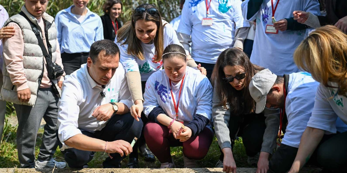 Bornova'da bir grup genç ve yetişkin, açık havada toprağa fidan dikmek veya tohum ekmek için bir araya gelmiş durumda. Beyaz tişörtler giymiş olan katılımcıların kıyafetlerinde el izleri bulunuyor ve boyunlarında kırmızı kurdeleli kimlik kartları asılı. Ön planda, beyaz gömlekli bir erkek ve yanında oturan down sendromlu bir genç kız, dikkatle toprağı inceliyor. Etraflarındaki diğer katılımcılar da yere çömelmiş veya ayakta durarak bu sürece eşlik ediyor; ortamda iş birliği ve dayanışma dolu bir atmosfer hakim.