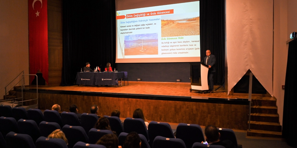 Bir konferans salonunda, kürsüde konuşma yapan bir kişi ve sahnede oturan iki panelist bulunmaktadır. Sahnenin arkasındaki dev ekranda 
