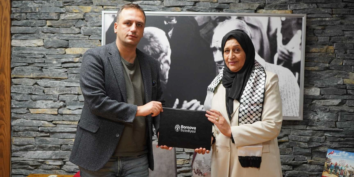 Bornova Belediye Başkanı Ömer Eşki ve Filistin'in Dura Belediyesi'nden bir temsilci, üzerinde Bornova Belediyesi logosu bulunan siyah bir dosyayı birlikte tutarak poz veriyor. Arka planda taş duvar üzerinde siyah beyaz bir Atatürk portresi yer alıyor.