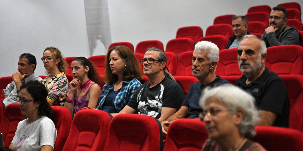 Konferans salonunda kırmızı koltuklarda oturan ve dikkatle sahneye bakan farklı yaş gruplarından oluşan bir izleyici grubu.