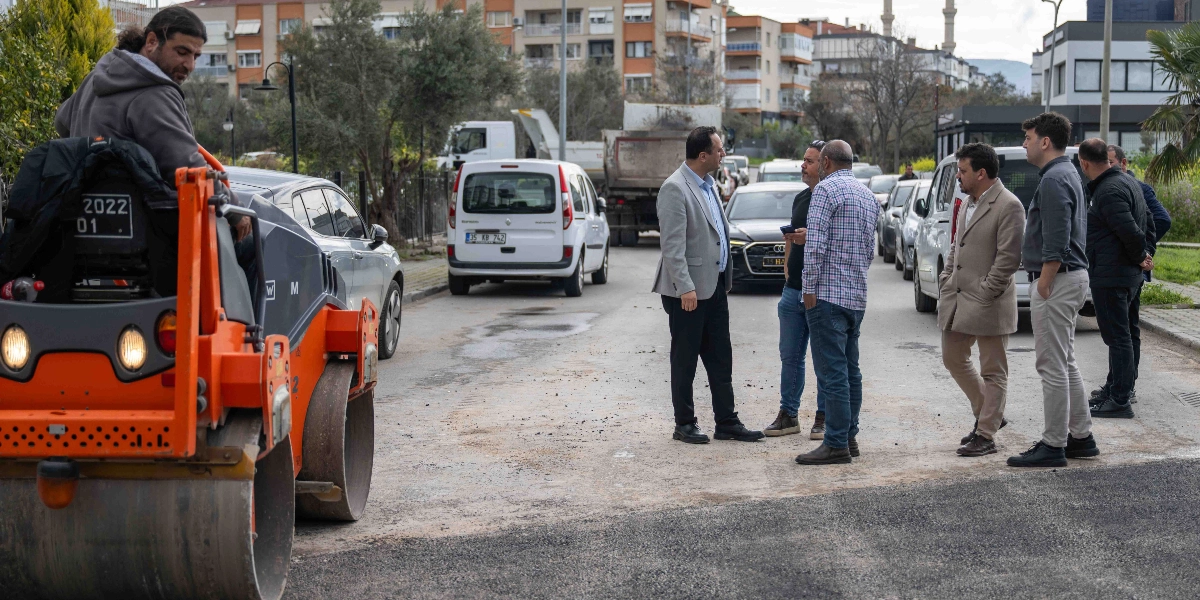 Turuncu bir silindir iş makinesinin operatörü ve yol çalışmasını inceleyen bir grup yetkili, asfaltlanmakta olan bir sokakta duruyor. Arka planda park halindeki araçlar, binalar ve ağaçlar görülüyor.