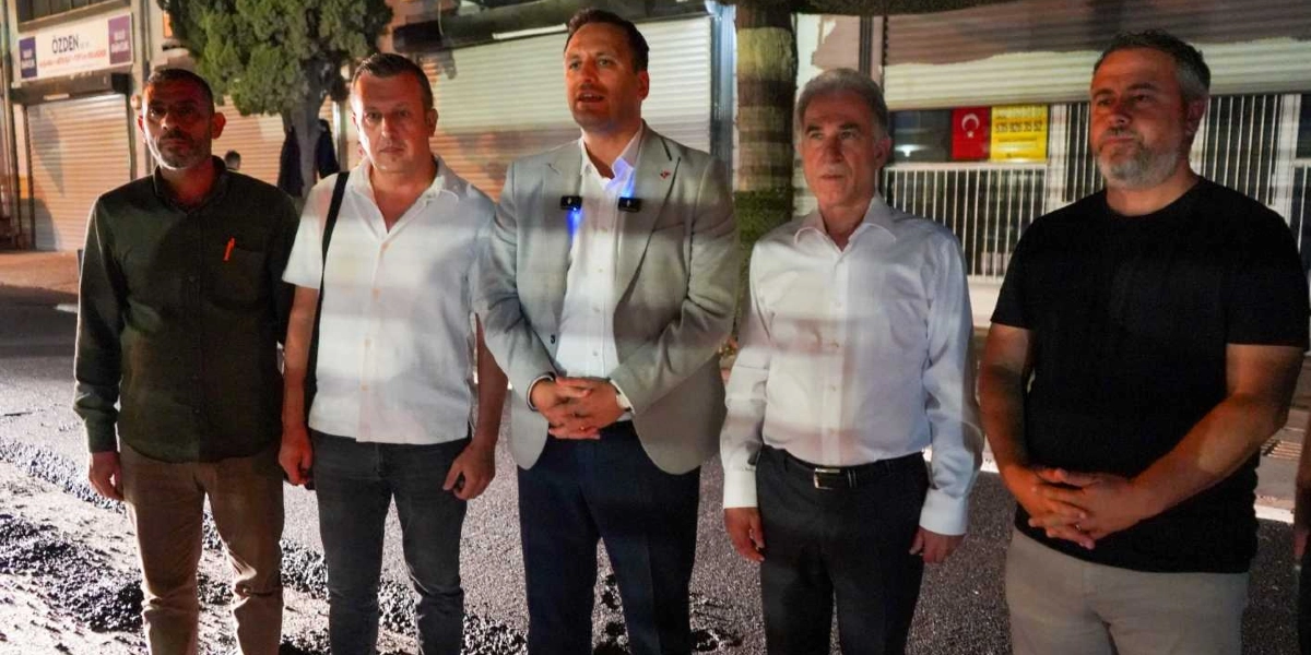 Gece vakti, yenilenmekte olan asfalt bir yolun önünde yan yana duran beş erkek. Ortada açık renk ceketli ve yakasında yaka mikrofonu bulunan bir kişi konuşma yaparken, diğer dört kişi onu dinliyor. Arka planda kepenkleri kapalı dükkanlar ve bir Türk bayrağı detayı görülüyor.