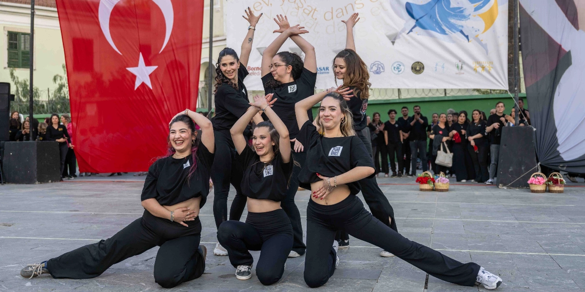 Açık havada, arkada büyük bir Türk bayrağı ve üzerinde dans temalı grafikler bulunan bir pankartın önünde, siyah tişört ve pantolon giymiş beş genç kadın dansçı koreografi sergiliyor. Dansçılardan üçü ön sırada, ikisi arka sırada yer alıyor; kollarını havaya kaldırmış veya vücutlarına yerleştirmiş şekilde dinamik bir poz veriyorlar. Arka planda etkinliği izleyen bir grup insan ve yerlerde çiçek sepetleri görülüyor.