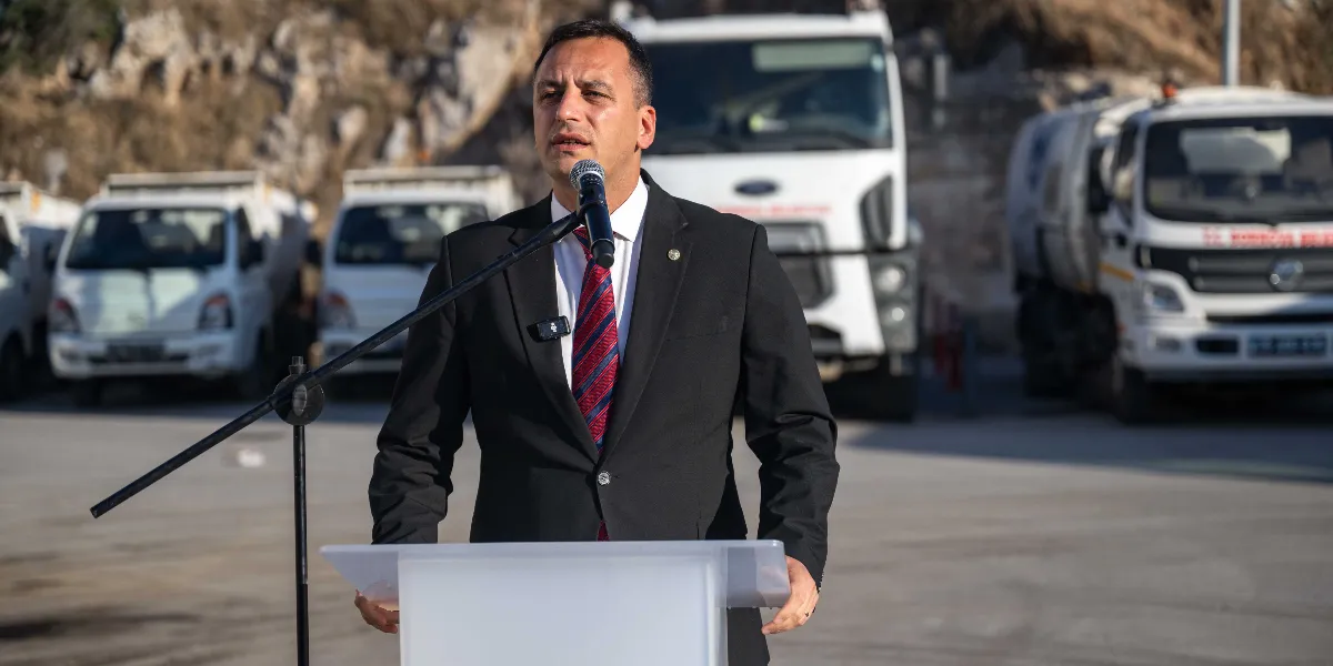 Siyah takım elbiseli ve kırmızı çizgili kravatlı bir erkek, açık havada, arkasında Bornova Belediyesi'ne ait beyaz hizmet araçlarının park halinde olduğu bir alanda kürsüde konuşma yapıyor.