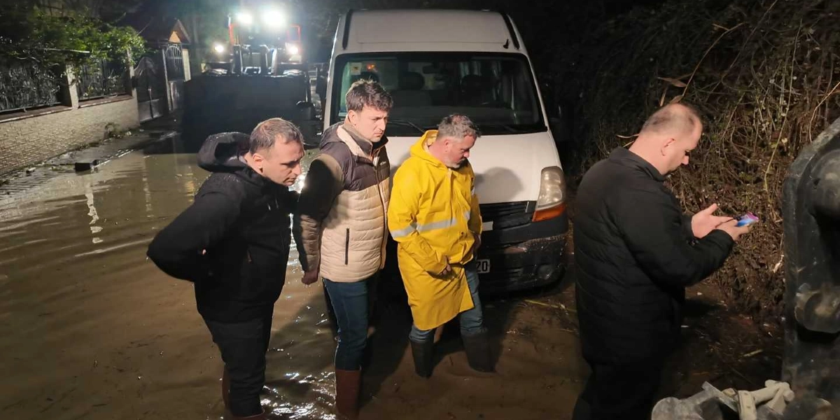 Gece vakti sel sularıyla kaplanmış bir sokakta, beyaz bir minibüsün önünde duran dört erkek, suyun seviyesini ve durumu incelemektedir. Sarı yağmurluklu ve çizmeli bir kişinin de aralarında bulunduğu grup, suyun içinde ayakta durmaktadır. Arka planda ise bir iş makinesinin farları yanmaktadır.