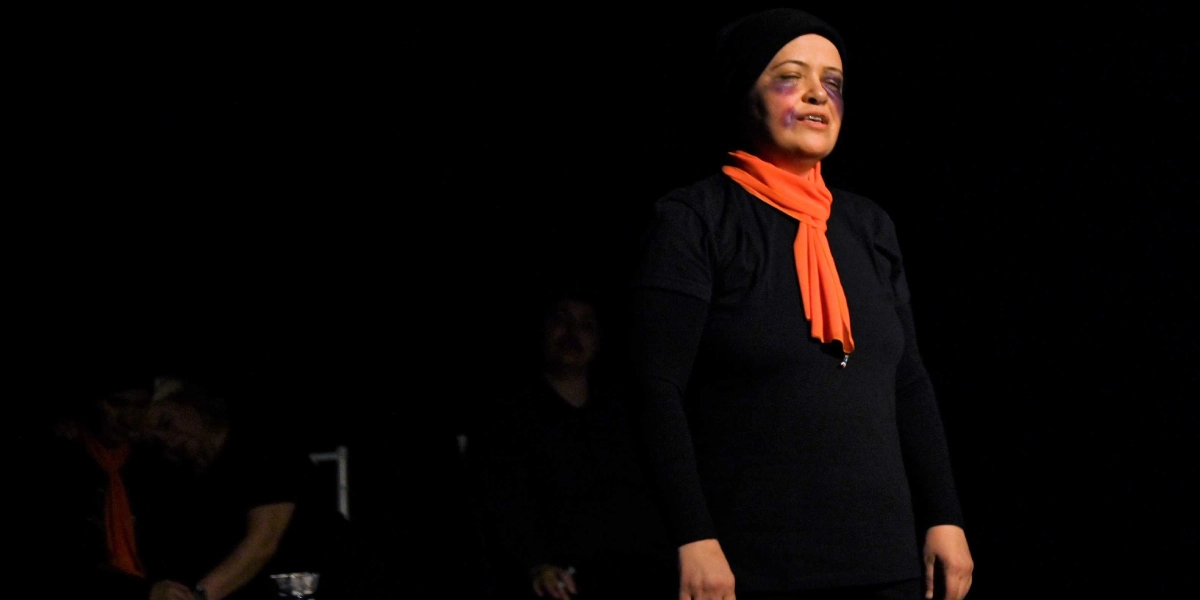 Siyah arka planlı bir sahnede, başında siyah bir bere, üzerinde siyah bir tişört ve boynunda turuncu bir fular bulunan bir kadın, göz çevresinde şiddet izlerini andıran mor makyajla performans sergiliyor. Kadının arkasında, karanlıkta silüetleri seçilen diğer oyuncular yer alıyor.