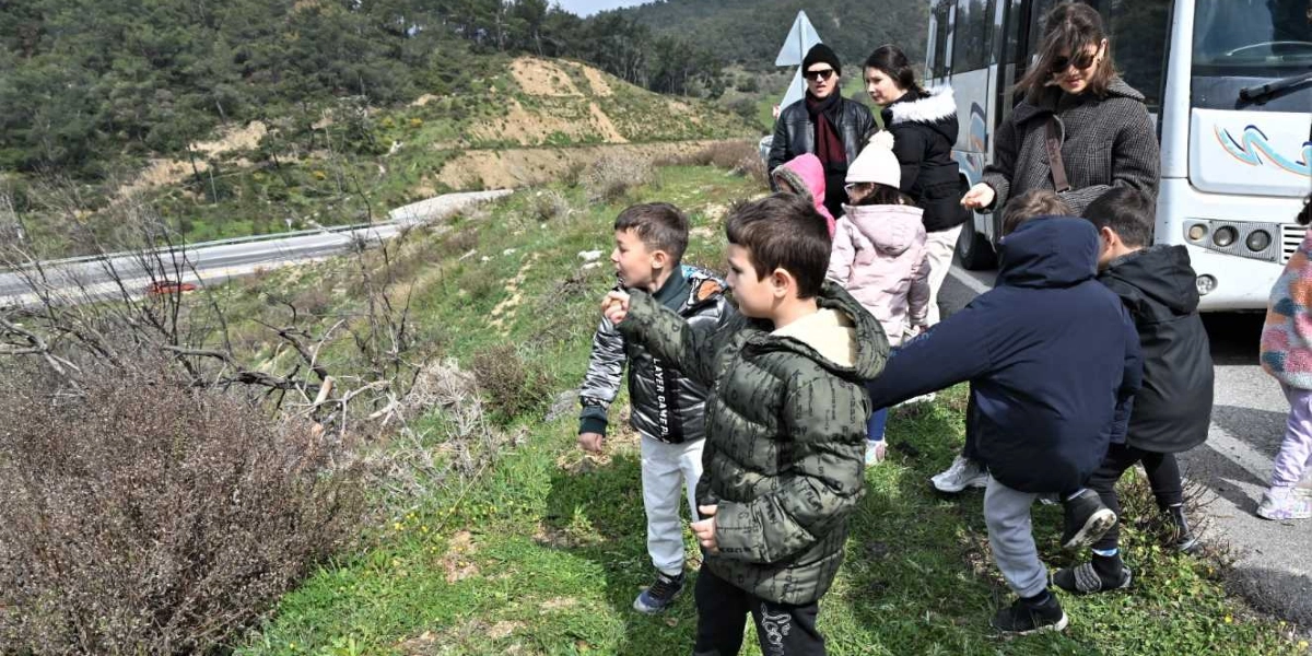 Açık havada, bir otobüsün yanında duran çocuklar ve onlara eşlik eden yetişkinler, yeşil bir yamaçta doğa etkinliği gerçekleştiriyor. Çocuklar kışlık montlar giymiş, ilgiyle çevrelerine bakıyor ve elleriyle bir şeyler işaret ediyor. Arka planda ağaçlıklı bir tepe ve aşağıdan geçen bir yol görülüyor.