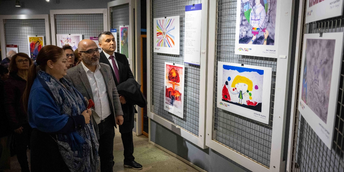 Bir sanat galerisinde, duvardaki metal panolara asılmış çocuk resimlerini inceleyen bir grup insan. Ön planda, koyu renkli kıyafetler giymiş bir kadın ve yanında gri ceketli, gözlüklü bir erkek ile takım elbiseli başka bir erkek, duvardaki resimlere dikkatle bakıyor. Panolarda, farklı tekniklerle yapılmış, üzerinde çocukların isimlerinin ve yaşlarının yazılı olduğu renkli resimler ve yanında Japonca metinler içeren bir pano sergileniyor.
