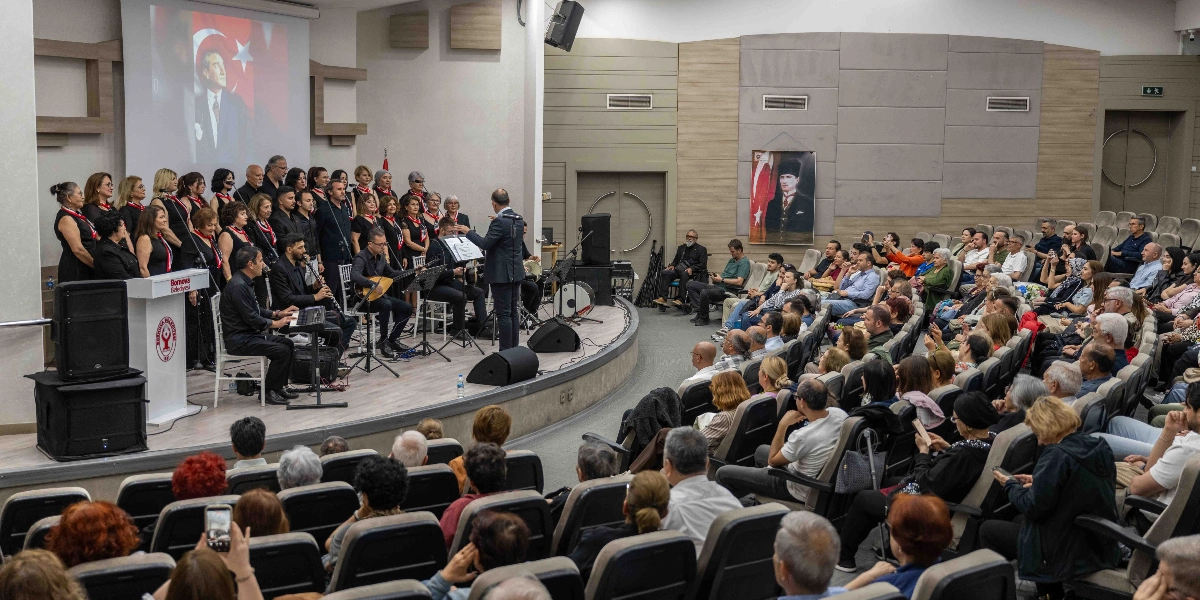 Modern bir konferans salonunda sahne alan bir koro ve orkestra, izleyicilere konser veriyor. Siyah kıyafetler giymiş koro üyeleri ve enstrüman çalan müzisyenler sahnedeki Bornova Belediyesi kürsüsünün arkasında yer alıyor. Sahnenin arka duvarındaki dev ekranda ve yan taraftaki panoda Mustafa Kemal Atatürk'ün portreleri bulunuyor. Salonun koltuklarında oturan kalabalık bir izleyici grubu, sahnedeki performansı ilgiyle takip ediyor.