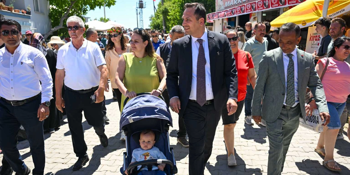 Açık havada, parke taşlı bir yolda yürüyen kalabalık bir grup insan. Grubun önünde, takım elbiseli ve kravatlı iki erkek ile bebek arabasını süren yeşil bluzlu bir kadın yer alıyor. Bebek arabasında oturan küçük bir çocuk etrafına bakıyor. Arka planda ise üzerinde 