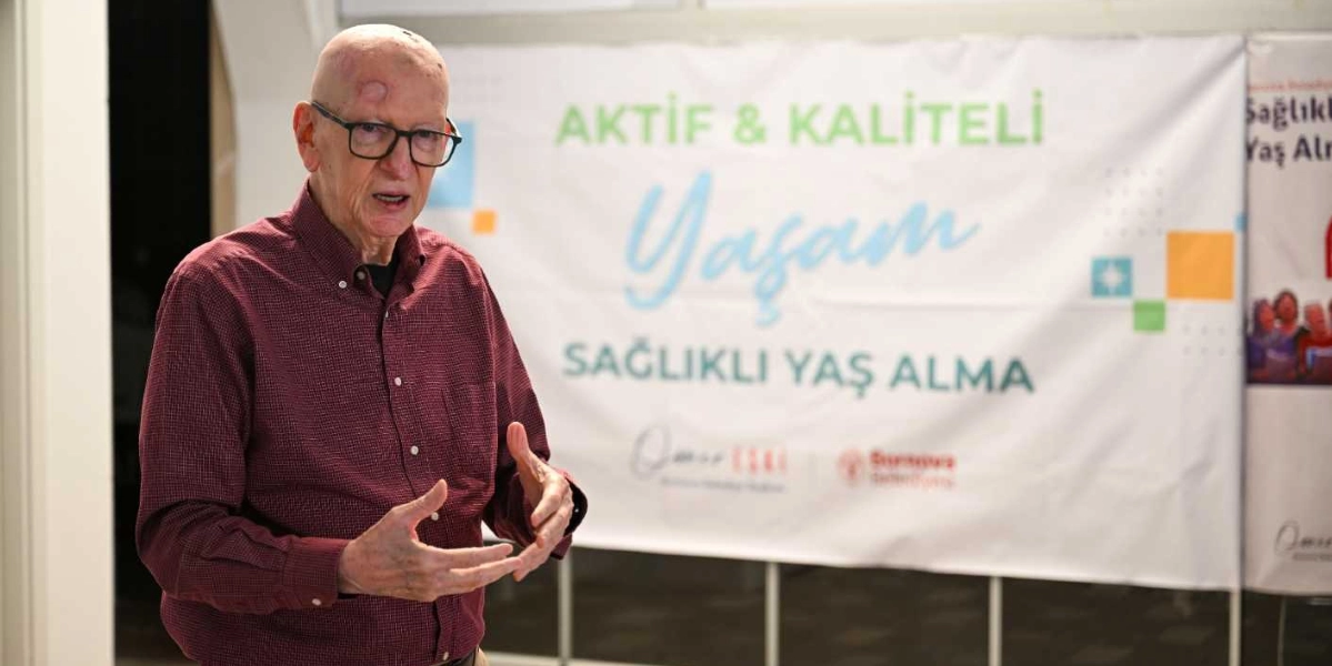 Gözlüklü, bordo gömlekli yaşlı bir adam, elleriyle konuşarak bir sunum yapıyor. Arkasında üzerinde 