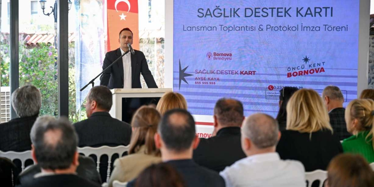 Kürsüde konuşma yapan bir erkek ve onu dinleyen katılımcıların yer aldığı bir toplantı salonu. Arkadaki büyük ekranda 