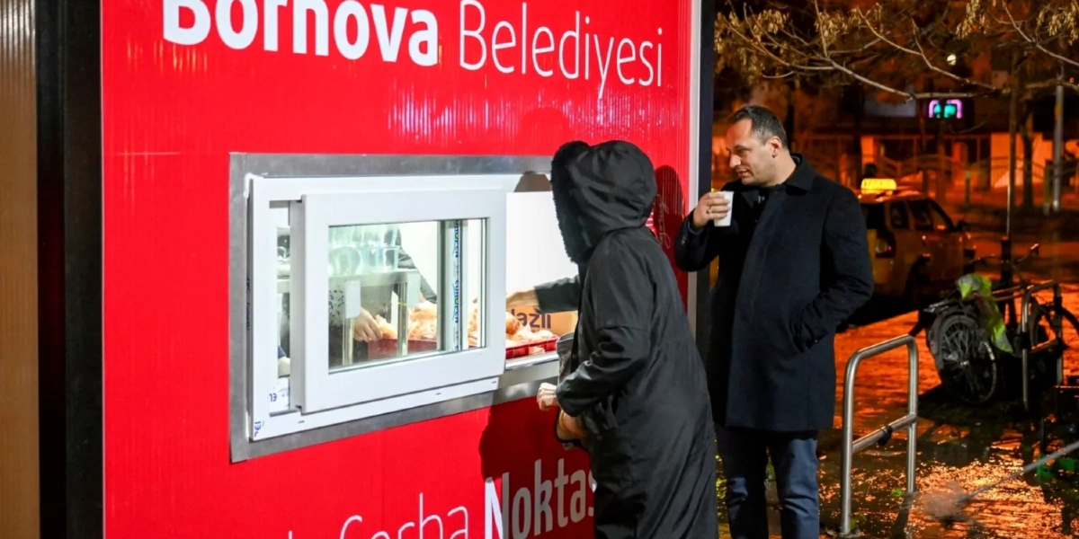 Üzerinde Bornova Belediyesi ve Çorba Noktası yazan kırmızı renkli bir stanttan, kapüşonlu bir kişi çorba alırken, yanında duran bir erkek elinde bir bardakla bekliyor.