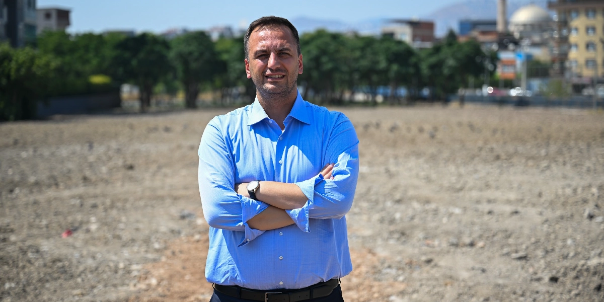 Mavi gömlekli, kısa saçlı ve hafif sakallı bir erkek, kolları göğsünde kavuşturulmuş şekilde, arkasında ağaçlık bir alan ve boş bir toprak arazinin bulunduğu bir dış mekanda kameraya gülümseyerek bakıyor.