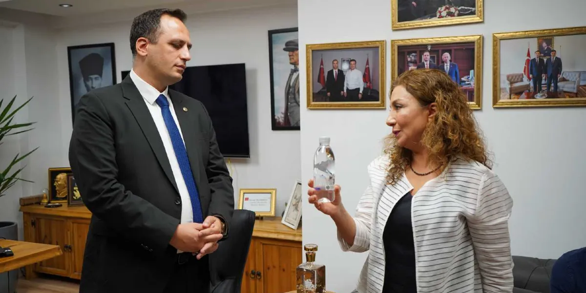 Koyu renk takım elbiseli ve mavi kravatlı bir erkek ile beyaz çizgili ceket giyen kıvırcık saçlı bir kadın, ofis ortamında ayakta durarak sohbet ediyor. Kadın, elinde tuttuğu şeffaf plastik bir su şişesini erkeğe gösteriyor. Arka planda duvarda Atatürk portreleri ve çeşitli etkinlik fotoğraflarının yer aldığı çerçeveler bulunuyor.