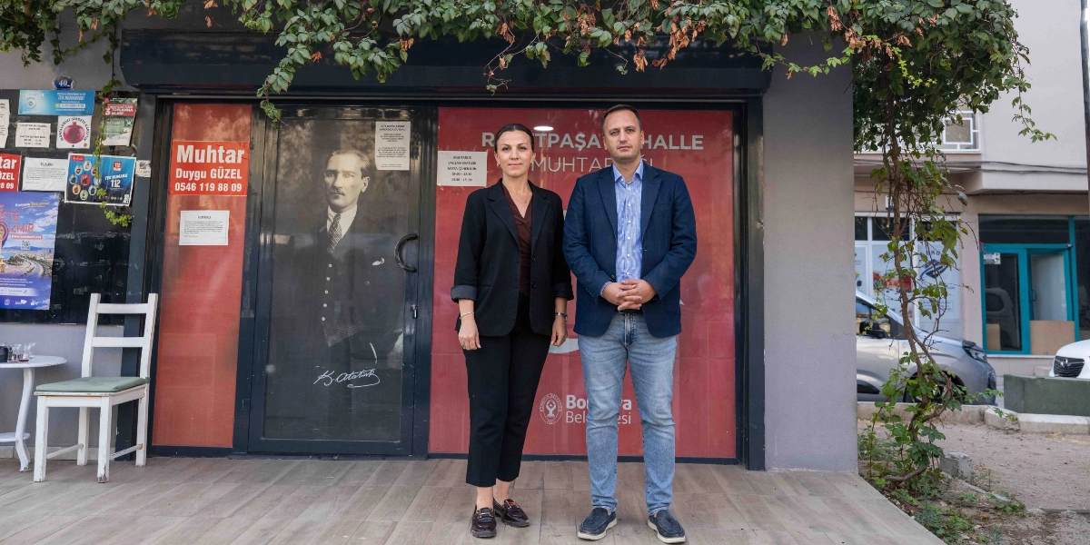 Bornova'da bulunan Reşitpaşa Mahalle Muhtarlığı binasının önünde, üzerinde Mustafa Kemal Atatürk'ün silüeti ve imzası bulunan cam kapının önünde duran bir kadın ve bir erkek. Kadın siyah ceket ve pantolon, erkek ise lacivert ceket ve kot pantolon giymiş şekilde kameraya bakıyor. Kapının üzerinde 