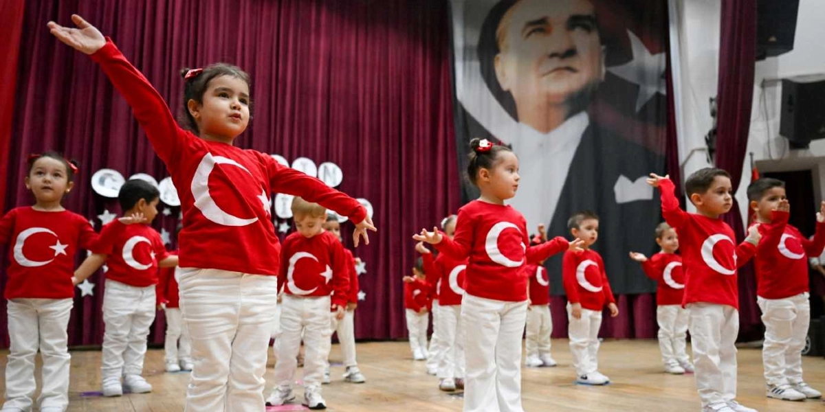 Kırmızı Türk bayraklı tişörtler ve beyaz pantolonlar giymiş anaokulu öğrencileri, bir sahne üzerinde dans gösterisi sergiliyor. Çocukların arkasında, bordo perdelerin önünde asılı duran büyük bir Mustafa Kemal Atatürk posteri yer alıyor. Sahne dekorunda ayrıca beyaz yıldız süslemeleri bulunuyor.