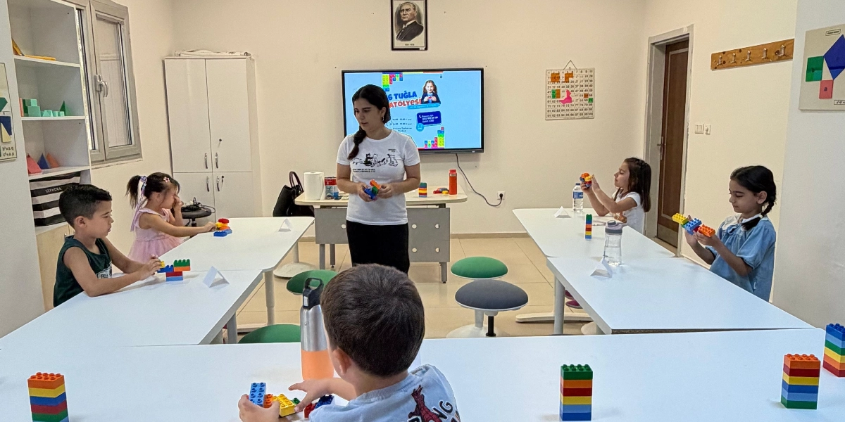 Sınıf ortamında beyaz masaların etrafında oturan çocuklar, önlerindeki renkli plastik tuğlalarla oyunlar oynuyor. Ayakta duran bir eğitmen, elindeki tuğlalarla çocuklara rehberlik ediyor. Arkadaki ekranda 6 Tuğla Atölyesi yazılı bir görsel yer alırken, duvarda Atatürk portresi ve sayı tablosu asılı bulunuyor.