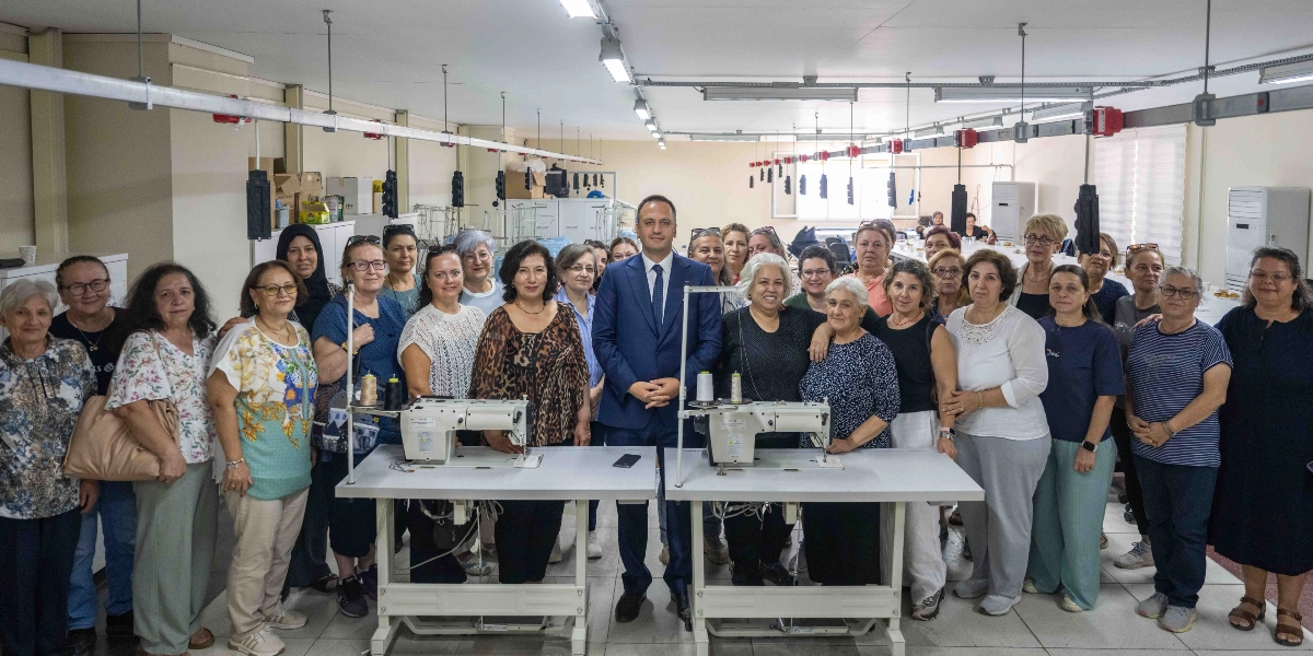 Geniş bir tekstil atölyesinde, aralarında takım elbiseli bir erkeğin de bulunduğu, çoğunluğu orta yaşlı kadınlardan oluşan kalabalık bir grup yan yana durarak poz veriyor. Grubun önünde iki adet sanayi tipi dikiş makinesi bulunuyor. Atölye ortamında tavandan sarkan aydınlatmalar ve çalışma masaları görülüyor.