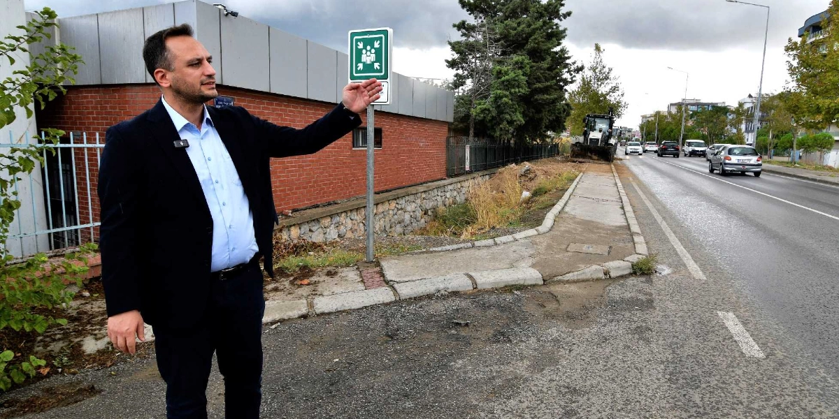 Koyu renk ceket ve açık mavi gömlek giymiş bir erkek, Bornova'da bir yol kenarında durarak eliyle yan taraftaki kaldırım ve yol çalışmasını işaret ediyor. Arkasında üzerinde toplanma alanı simgesi bulunan bir tabela, kırmızı tuğlalı bir bina duvarı ve ileride iş makinesinin bulunduğu bir cadde yer alıyor.