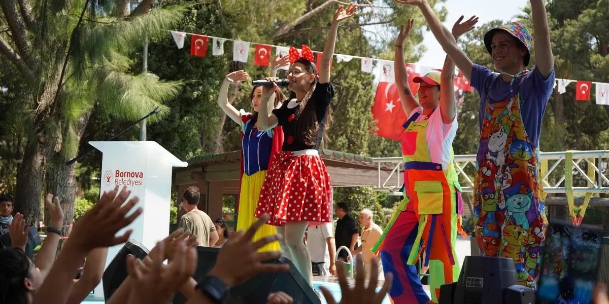 Açık havada düzenlenen çocuk şenliğinde, üzerinde Bornova Belediyesi logosu bulunan bir kürsünün yanında, kostümlü üç animatör sahne alıyor. Minnie Mouse kostümlü bir kadın mikrofonla şarkı söylerken, yanında Pamuk Prenses kostümlü bir kadın ve renkli, kedi desenli tulum giymiş bir genç, kolları havada çocukları eğlendiriyor. Arka planda Türk bayrakları ve ağaçlar görülürken, ön planda sahneyi izleyen çocukların havaya kaldırdığı elleri seçiliyor.