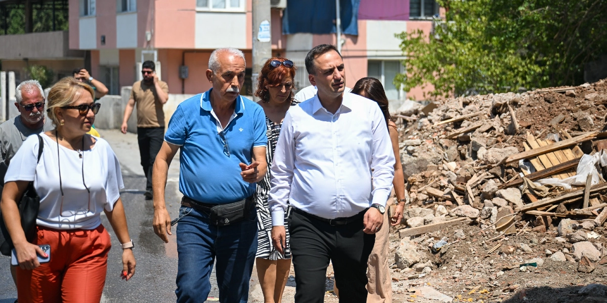 Beyaz gömlekli bir yetkili ve beraberindeki heyet, yıkım çalışması yapılan bir alanda incelemelerde bulunuyor. Mavi polo yaka tişörtlü bir kişi, el hareketleriyle çevreyi işaret ederek yanındaki yetkiliye bilgi veriyor. Arka planda ise moloz yığınları ve çok katlı bir bina görülüyor.