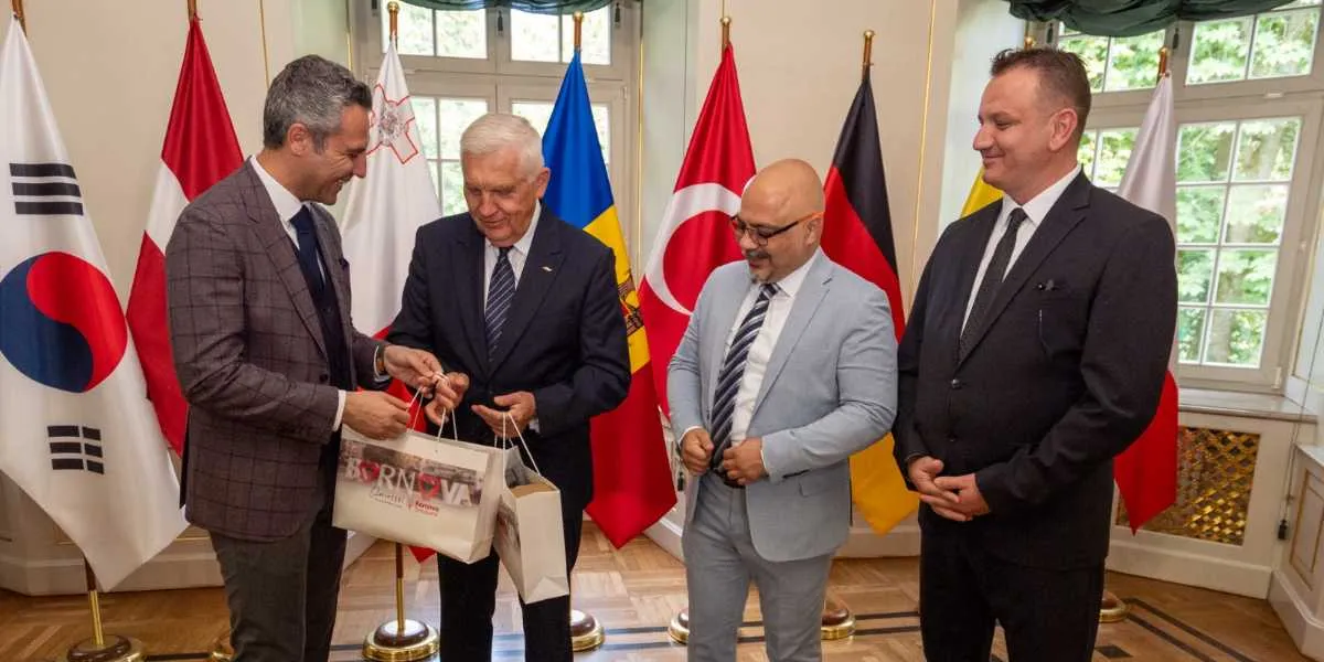 Resmi bir ortamda, arkalarında çeşitli ülkelerin bayraklarının bulunduğu bir odada, dört erkek yan yana duruyor. Soldaki kişi, üzerinde 