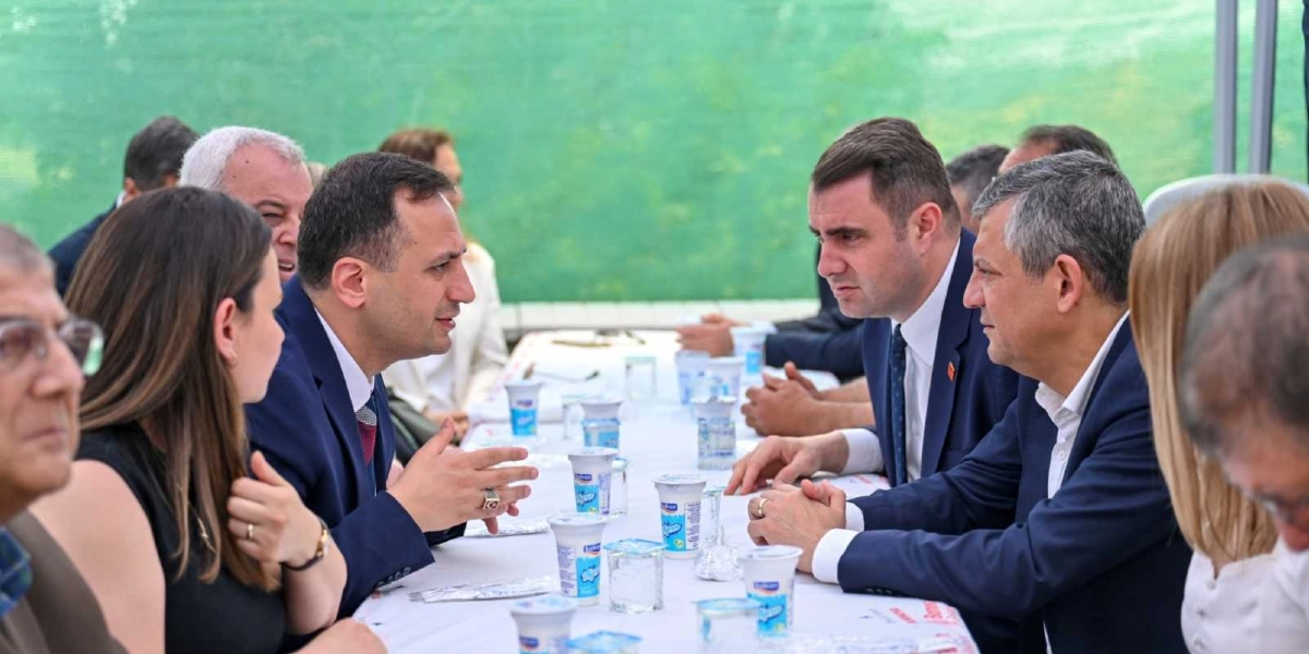 CHP Genel Başkanı Özgür Özel, Bornova'daki bir etkinlikte masada oturan diğer yetkililerle sohbet ediyor. Beyaz örtülü uzun bir masanın etrafında toplanan katılımcıların önünde küçük paketlerde ayranlar bulunuyor. Özgür Özel, masanın sağ tarafında koyu renkli bir takım elbise içinde, karşısındaki kişiyle konuşurken görülüyor.