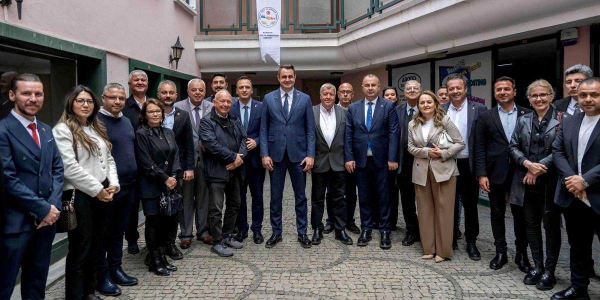 Bornova Balkan Göçmenleri Derneği binasının avlusunda, aralarında CHP İzmir Milletvekili ve yerel yöneticilerin bulunduğu, çoğunluğu takım elbiseli erkekler ve şık giyimli kadınlardan oluşan yaklaşık yirmi kişilik bir grup, objektiflere poz veriyor. Grubun merkezinde lacivert çizgili takım elbisesiyle bir erkek yer alırken, arkadaki duvarda derneğin logosunun bulunduğu bir flama asılı duruyor.