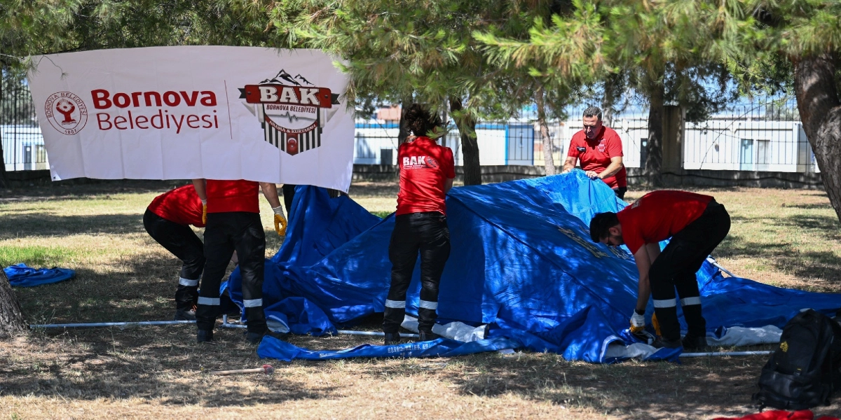 Ağaçlık bir alanda, üzerinde Bornova Belediyesi ve BAK Arama Kurtarma logolarının bulunduğu beyaz bir pankartın altında, kırmızı tişörtlü ve siyah pantolonlu arama kurtarma ekibi üyeleri mavi bir çadır kuruyor.