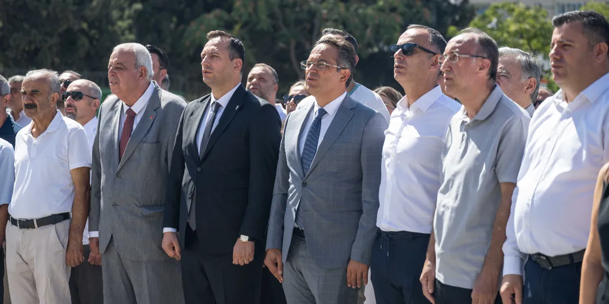 Açık havada, yan yana duran ve ciddi bir ifadeyle karşıya bakan bir grup erkek siyasetçi. Önde, takım elbiseli ve kravatlı olanlar ile beyaz gömlekli ve güneş gözlüklü kişilerin yer aldığı grup, resmi bir tören veya anma etkinliği sırasında saygı duruşunda bulunuyor.