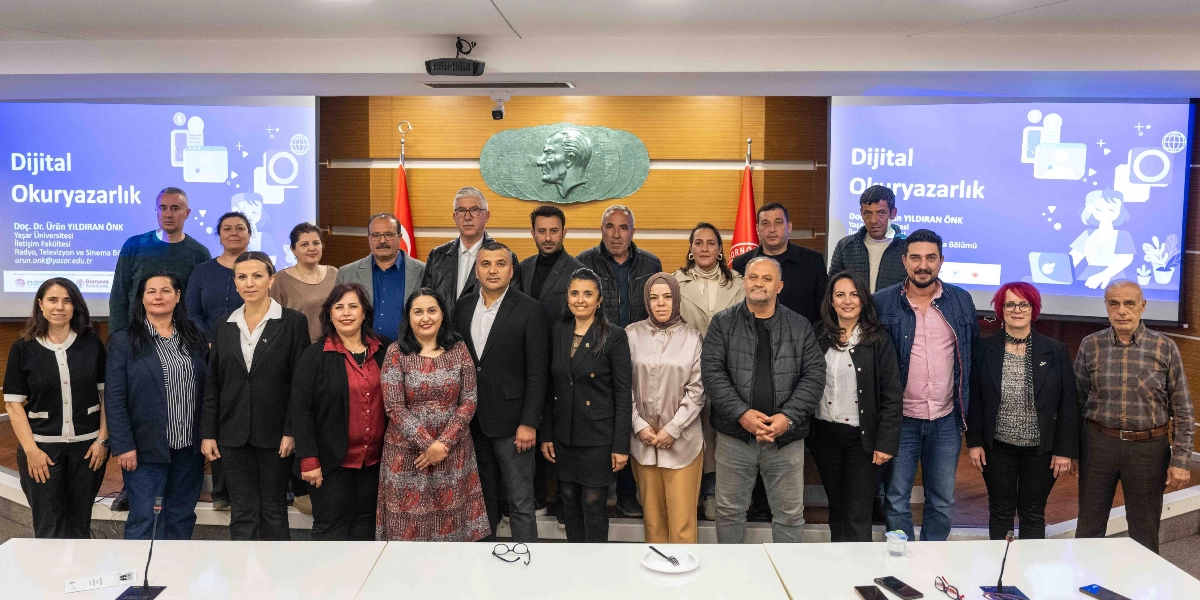 Bornova Muhtarlar Akademisi kapsamında düzenlenen Dijital Okuryazarlık eğitimi sonrasında, katılımcı muhtarlar ve eğitmen Doç. Dr. Ürün Yıldıran Önk, arkalarında Atatürk silüeti bulunan bir toplantı salonunda toplu halde poz veriyor. Arka planda yer alan iki büyük ekranda 
