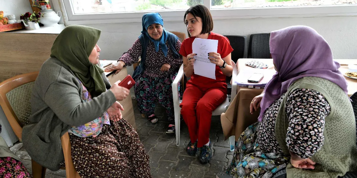 Kırmızı üniformalı genç bir sağlık çalışanı, köy ortamında üç kadına elindeki sağlık bilgilendirme broşürü üzerinden eğitim veriyor. Kadınlar dikkatle sağlık çalışanını dinlerken, sağlık çalışanı elindeki kağıdı göstererek açıklamalarda bulunuyor.