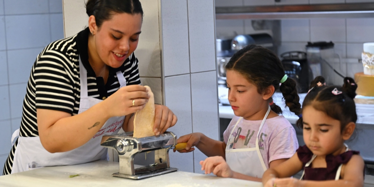 Beyaz önlüklü genç bir kadın, iki küçük kız çocuğuna mutfak tezgahı üzerinde metal bir makarna makinesini nasıl kullanacaklarını gösteriyor. Kadın hamuru makineye yerleştirirken, yanındaki kız çocuğu makinenin kolunu çevirerek ona yardımcı oluyor, diğer çocuk ise onları dikkatle izliyor. Hepsi mutfak önlüğü takmış şekilde profesyonel bir mutfak ortamında çalışıyor.