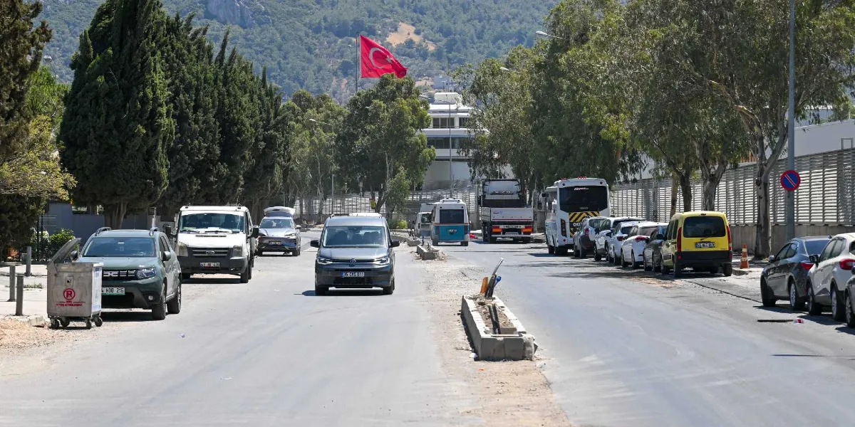 Ağaçlarla çevrili Pınar Caddesi'nde devam eden yol çalışmaları sırasında, trafiğin aktığı asfalt yolun ortasında beton bariyerler ve kenarlarda park halindeki araçlar görülüyor. Arka planda tepelik bir alan üzerinde dalgalanan Türk bayrağı yer alıyor.