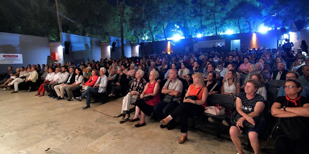 Gece vakti açık hava tiyatrosunda düzenlenen bir konseri izleyen kalabalık bir seyirci grubu. Sahneye doğru dönük şekilde sıralanmış koltuklarda oturan farklı yaş gruplarından insanlar, dikkatle sahneye bakıyor. Arka planda ağaçlar ve sahneyi aydınlatan mavi ışıklar görülüyor. Sol taraftaki duvarda üzerinde yazı bulunan beyaz bir bilgilendirme panosu yer alıyor.