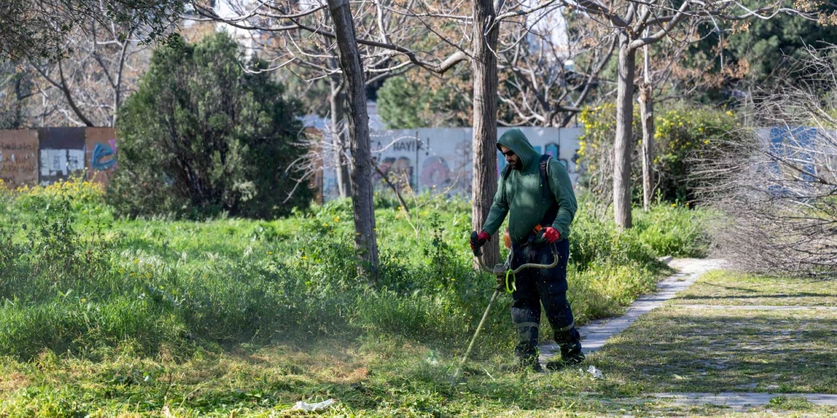 Yeşil kapüşonlu sweatshirt, güneş gözlüğü ve kırmızı eldiven giyen bir görevli, parktaki uzun otları motorlu tırpanla biçiyor. Arka planda ağaçlar ve üzerinde grafiti bulunan bir duvar yer alıyor.
