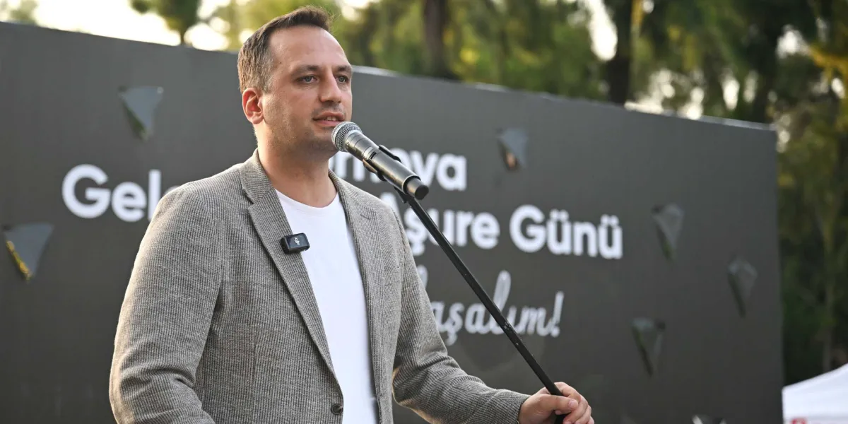 Beyaz tişört ve gri ceket giymiş bir erkek, önündeki mikrofona konuşma yapıyor. Arka planda üzerinde 