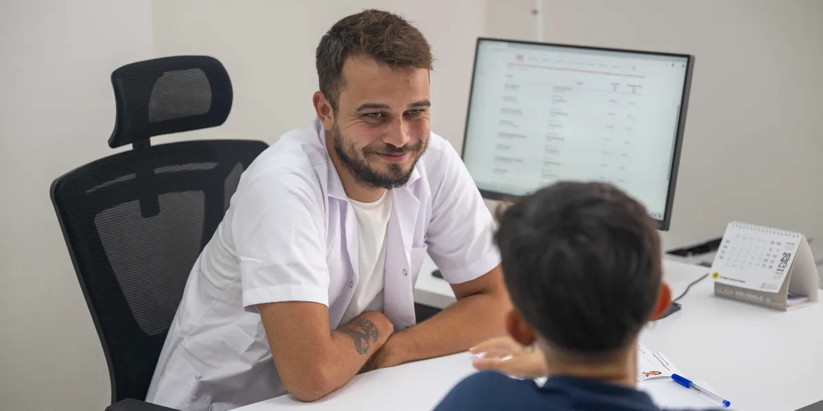 Beyaz önlüklü, sakallı genç bir uzman, ofis ortamında masanın karşısında oturan bir çocukla gülümseyerek ilgileniyor. Masanın üzerinde bir bilgisayar ekranı ve 2025 yılına ait bir masa takvimi bulunuyor.