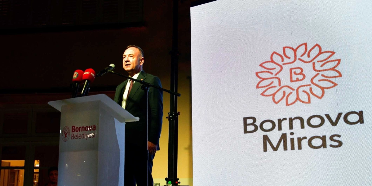 Koyu renk takım elbiseli bir konuşmacı, üzerinde Bornova Belediyesi logosu bulunan bir kürsüde mikrofon başında konuşma yapıyor. Konuşmacının sağ tarafında yer alan büyük ekranda, stilize edilmiş bir çiçek motifinin ortasında 